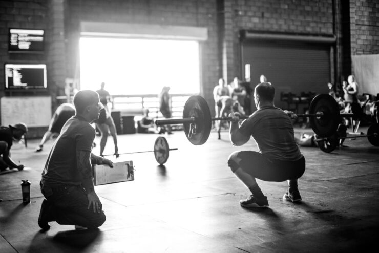 Trainer observing cross training athlete doing squats with barbell in gym